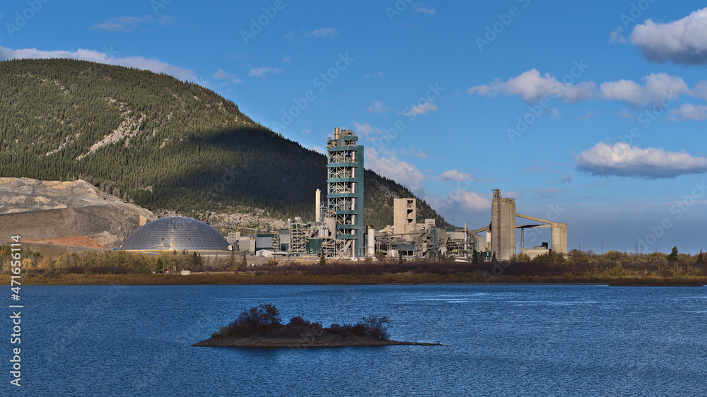 View of huge industrial cement plant in Bow Valley near Canmore, Alberta, Canada in the Rocky Mountains in autumn season with lake in front.
