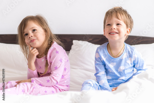 Two Preschool Toddler Children Siblings Boy And Girl Smiling Look At Side In Pink Blue Pajamas On White Bed. Little Baby Twins Have Fun. Happy Kids On Quarantine At Home. Friendship, Family Concept