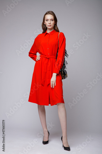 High fashion photo of a beautiful elegant young woman in a pretty red dress, black handbag posing over white, soft gray background. Studio Shot.