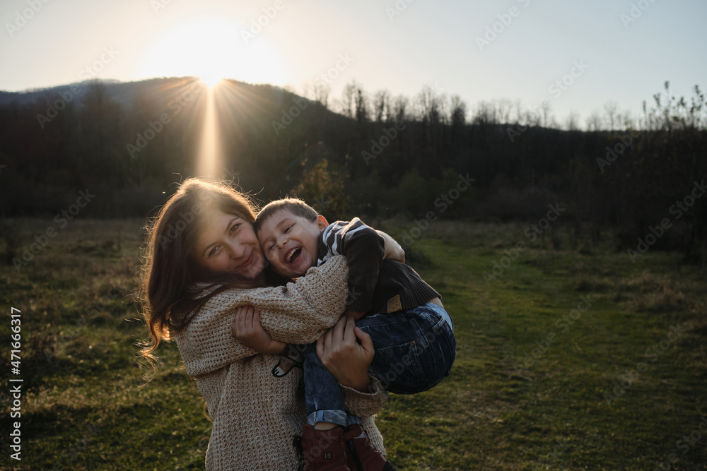 Spend childhood in nature with beloved mother. Joy of motherhood. Warm setting sun. Caucasian boy in striped sweater is sitting merrily in arms of woman and they are both laughing hugging. Spend childhood in nature with beloved mother. Joy of motherhood. Warm setting sun. Caucasian boy in striped sweater is sitting merrily in arms of woman and they are both laughing hugging.