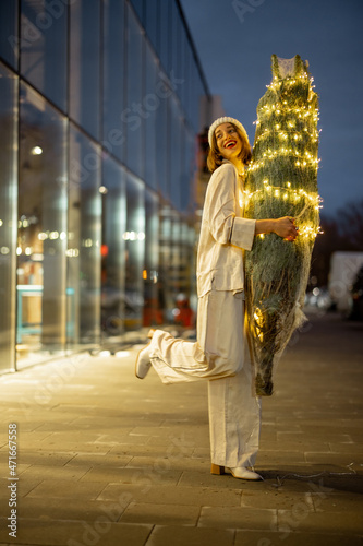 Portrait of a woman in pajamas and winter hat with illuminated Christmas tree near the shopping centre at dusk. Concept of preparation for the winter holidays. Idea of a winter fairytale and mood photo