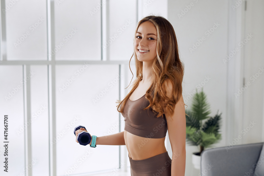 A slim actress is warming up with dumbbell while standing on the mat and smiling at the camera.