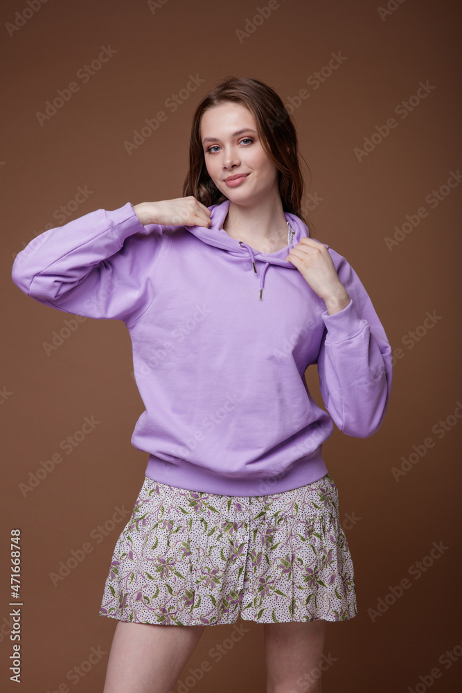Obraz premium High fashion photo of a beautiful elegant young woman in a pretty short beige shirt dress with floral patterns, white sneakers, purple sweatshirt posing over brown background. Studio Shot. Portrait