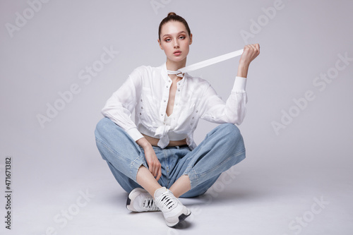 High fashion photo of a beautiful elegant young woman in a pretty white shirt and sneakers, blue denim jeans posing over white, soft gray background. Studio Shot. The model is sitting