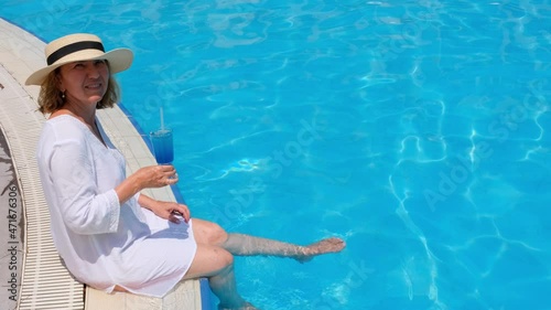 relaxing senior woman dangling her legs in an outdoor swimming pool holding a blue refreshing cocktail wearing a straw hat. Summer rest. All inclusive