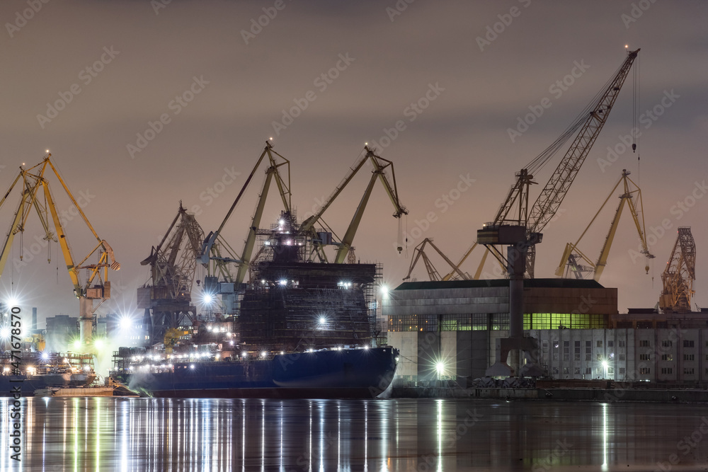 The construction of nuclear icebreakers at night, cranes of of the Baltic shipyard in a frosty winter day, steam over the Neva river, smooth surface of the river, fog