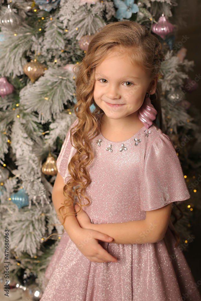 a large portrait of a beautiful little girl in a festive pink dress stands at the Christmas tree