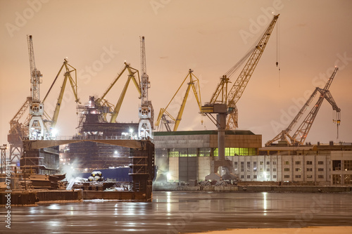 The construction of nuclear icebreakers at night, cranes of of the Baltic shipyard in a frosty winter day, steam over the Neva river, smooth surface of the river, fog