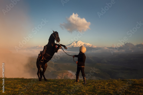 An epic view of the largest mountain in Europe, Elbrus. In the foreground is a Caucasian man in a fur hat and a rearing horse. Mountain horse of the Karachai breed. Bermamyt plateau. North Caucasus.