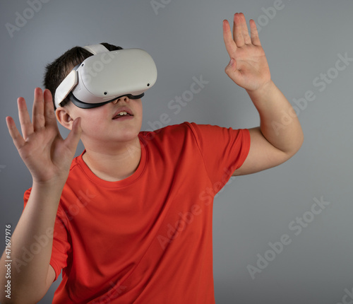 A teenager with glasses-viar looks up and raised his hands while playing a game. A boy in a red T-shirt on a gray background. Virtual reality