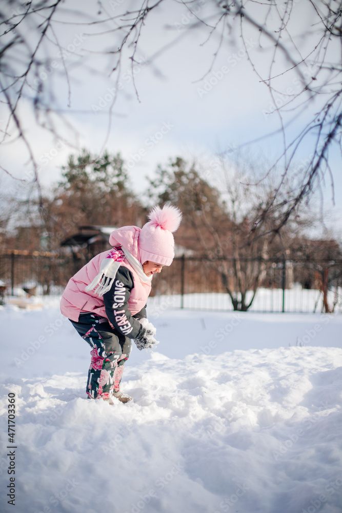 Child is playing in nature. Little girl in warm suit plays in backyard and enjoys sunny day in winter, she stands on snowdrift and plays snowballs, side view. Child is playing in nature. Little girl in warm suit plays in backyard and enjoys sunny day in winter, she stands on snowdrift and plays snowballs, side view.