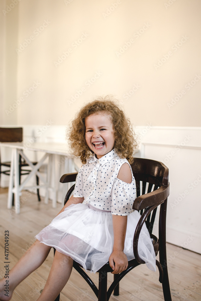 a beautiful little curly-haired girl sitting in the middle of a large room on a chair. smiling happily a beautiful little curly-haired girl sitting in the middle of a large room on a chair. smiling happily