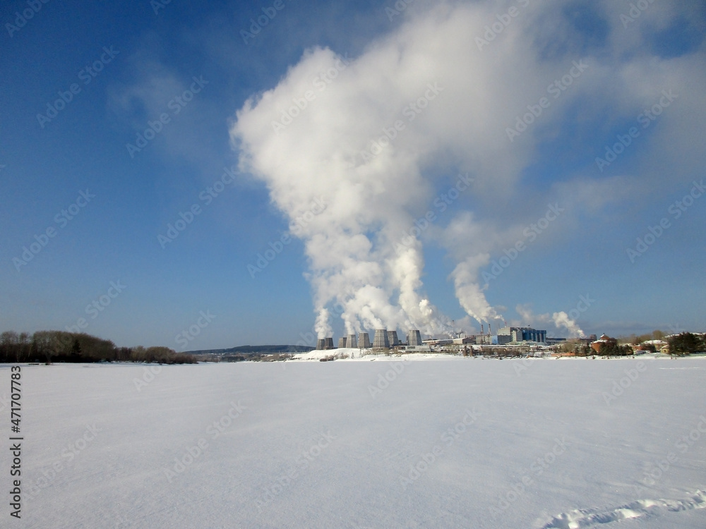 The winter open space of a frozen urban pond with beautiful white clouds of vapor from the cooling towers of the plant in the background, blue sky and white snow cover on a bright sunny day