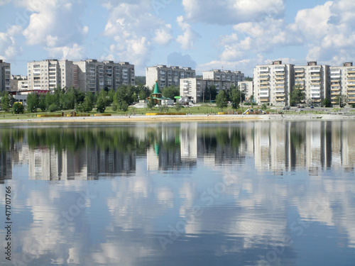 The colorful sunny view of the embankment of the Russian town with an entertainment center and multi-storey buildings is beautifully reflected in the calm water along with white clouds in the blue sky
