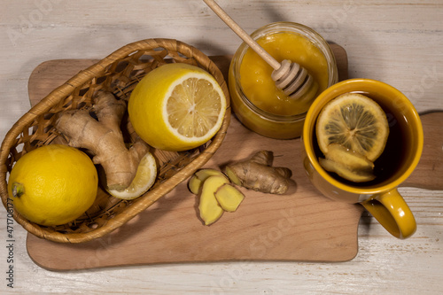  On a cutting board is a cup of tea with lemon, a jar of honey, lemons and ginger.