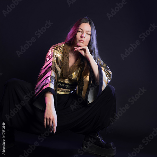 Stylish and daring image, futuristic fashion. A young woman in a multicultural outfit mixing Asian and European style, a golden kimono shirt and trousers, photo in the studio on black