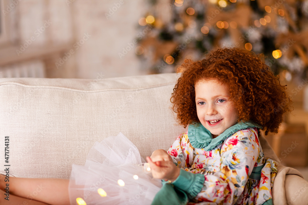 Pretty little red hair girl having fun with garland near Christmas tree waiting for miracle. Holiday mood, lights on background, cozy home atmosphere