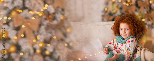 Pretty little red hair girl having fun with garland near Christmas tree waiting for miracle. Holiday mood, lights on background, cozy home atmosphere