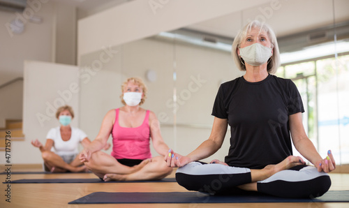 Group of aged ladies in face masks doing lotus pose on rugs in fitness room.