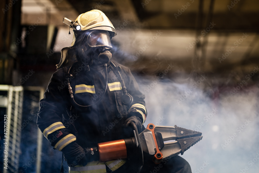 Fototapeta premium firefighter portrait wearing full equipment, oxygen mask, and power hydraulic cutting tool, smoke and fire trucks in the background.