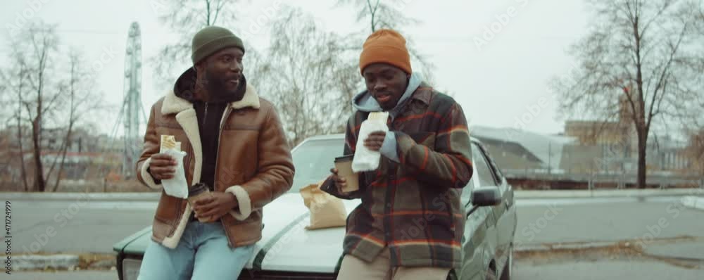 Two African American men leaning on car, eating sandwiches, drinking coffee from to go cups and speaking