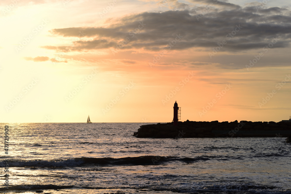 In the evening, at sunset, there is a lighthouse on the horizon. In the background a small sailing boat sailing across the sea.