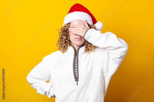 Curly young woman in a santa hat with palms closed her eyes on a yellow background. © Alex