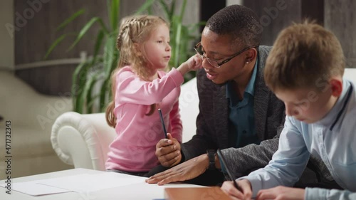 Blonde preschooler girl pupil touches black man teacher ear learning body parts names with brother at private English lesson at home slow motion