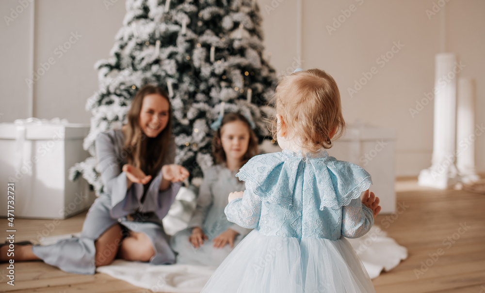 a little daughter stands with her back and takes the first steps to her mother and sister in a room with a Christmas tree