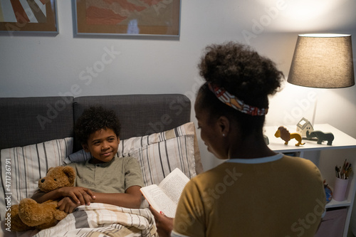 Blackwoman with curly hair sitting in nursery room and reading fairy tale to little son lying with toy in bed