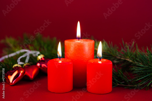 Scena natalizia con tre candele rosse accese e con addobbi natalizi a forma di cuore circondati da rami di abete su sfondo rosso. Natale. Auguri. Dicembre. Feste invernali.