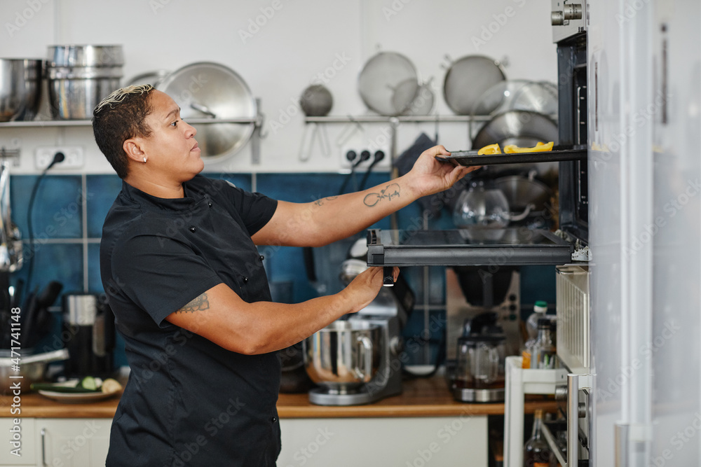 Side view portrait of tattooed female chef opening oven in professional kitchen, copy space Side view portrait of tattooed female chef opening oven in professional kitchen, copy space