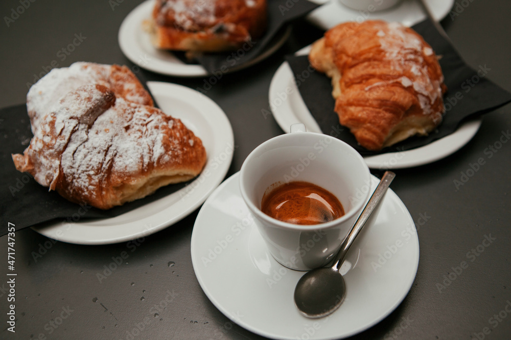 The table with sweets (pastries: croissant, cookies), white cups (mug) with hot black coffee (espresso), the saucer, the spoons at a local bar. Traditional Italian breakfast. Milan, Lombardy, Italy The table with sweets (pastries: croissant, cookies), white cups (mug) with hot black coffee (espresso), the saucer, the spoons at a local bar. Traditional Italian breakfast. Milan, Lombardy, Italy