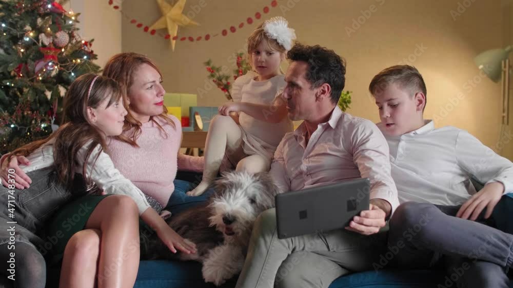 A happy family is sitting on the couch in living room and having a video call via tablet. The room is decorated in the Christmas spirit. Also, their dog is on the couch with them. A happy family is sitting on the couch in living room and having a video call via tablet. The room is decorated in the Christmas spirit. Also, their dog is on the couch with them.