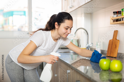 Attractive smiling young woman cleaning kitchen counter with spray bottle and rag