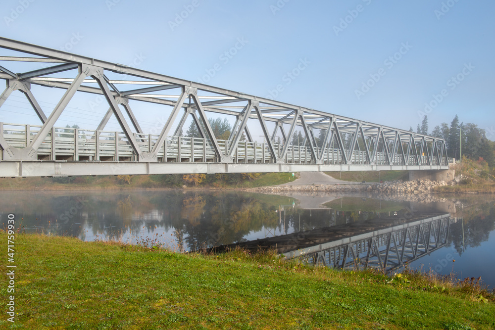 A steel girder bridge spans across a narrow river under blue skies and clouds. The deck of the bridge is pavement and there's a guide rail on both sides. The metal bridge is used for vehicle travel.