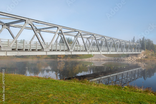 Fototapeta Naklejka Na Ścianę i Meble -  A steel girder bridge spans across a narrow river under blue skies and clouds. The deck of the bridge is pavement and there's a guide rail on both sides. The metal bridge is used for vehicle travel.