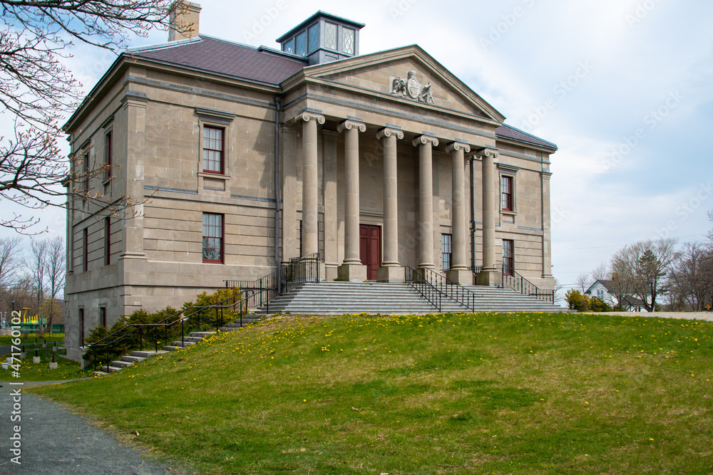 The exterior facade of a historic government building with large vintage marble pillars or columns, red door, blue sky, trees and a brick entrance leading up steps to the large antique stone building.