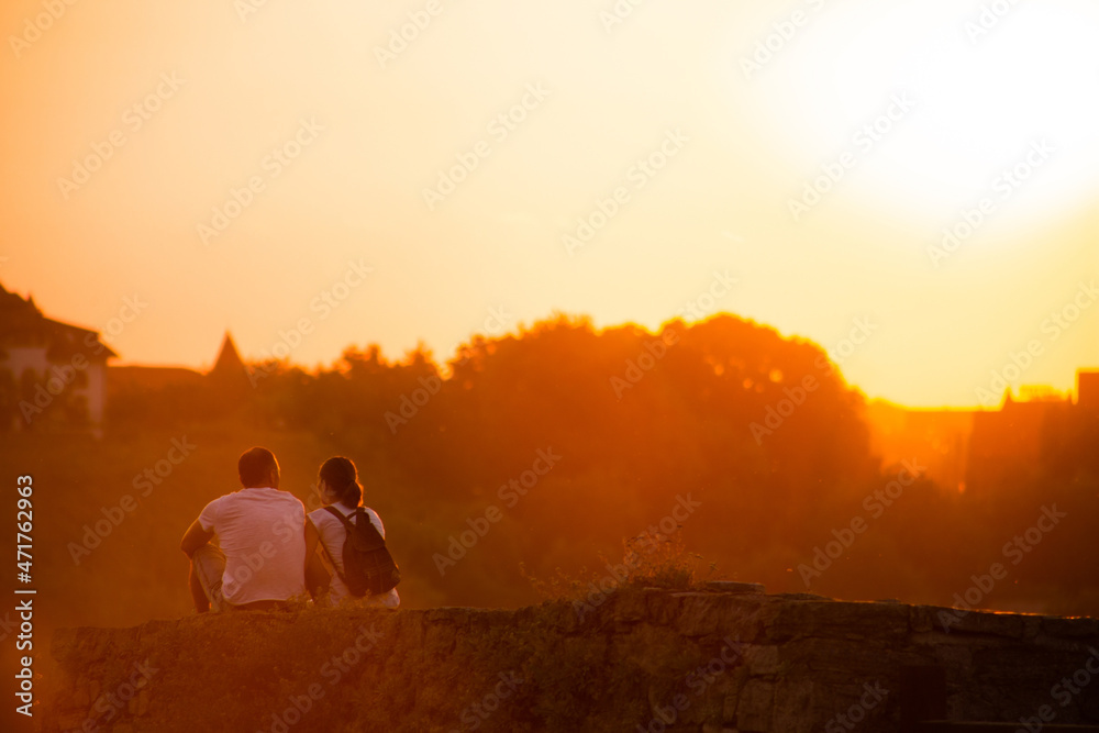 A couple of young people sit on the edge of a cliff as the sunset.