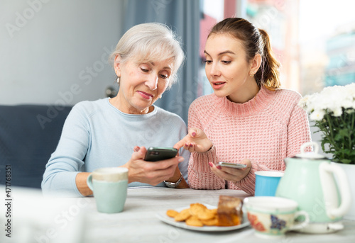 Positive young girl showing pictures on her smartphone to interested elderly mother while sitting at table in cozy dining room.