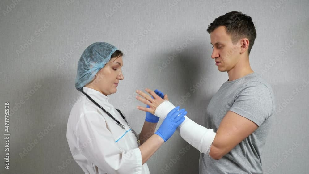 Brunette doctor in medical cap and gloves examines broken spoke bone in gypsum of young short-haired man wearing grey T-shirt in clinic. Brunette doctor in medical cap and gloves examines broken spoke bone in gypsum of young short-haired man wearing grey T-shirt in clinic.