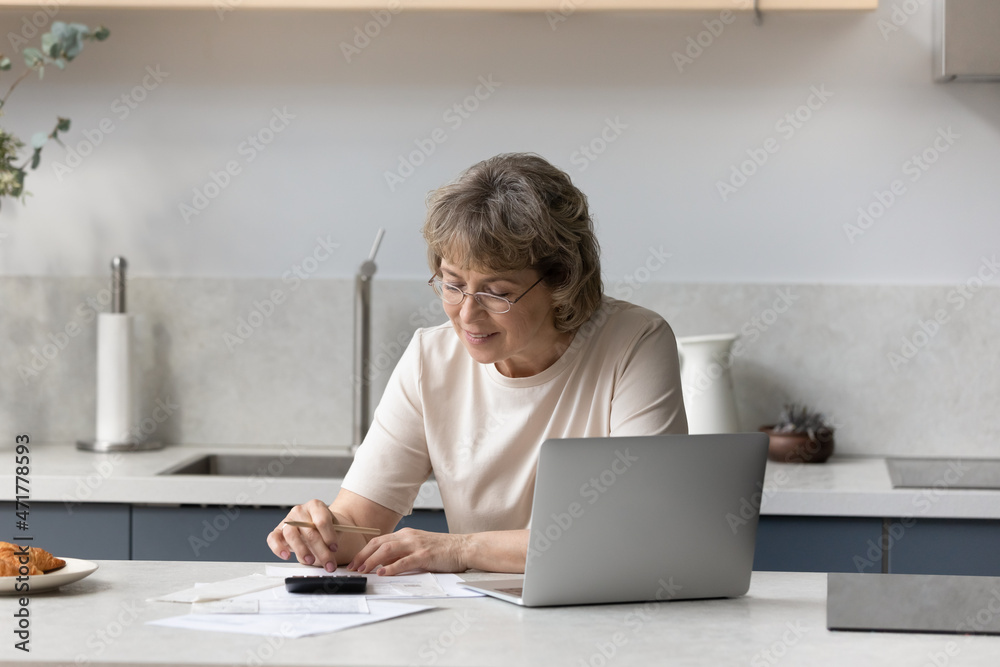 Focused mature homeowner calculating taxes, insurance fees, counting budget, expenses, using calculator, online financial app on laptop, doing paperwork, checking paper bills, invoices Focused mature homeowner calculating taxes, insurance fees, counting budget, expenses, using calculator, online financial app on laptop, doing paperwork, checking paper bills, invoices