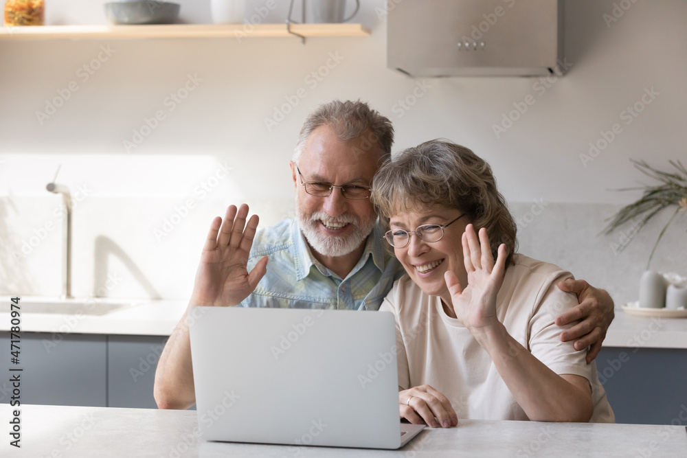 Obraz premium Happy senior couple talking to children on video call, waving hello at laptop screen, smiling, laughing, sitting at kitchen table. Grandparents enjoying remote communication with family, using laptop