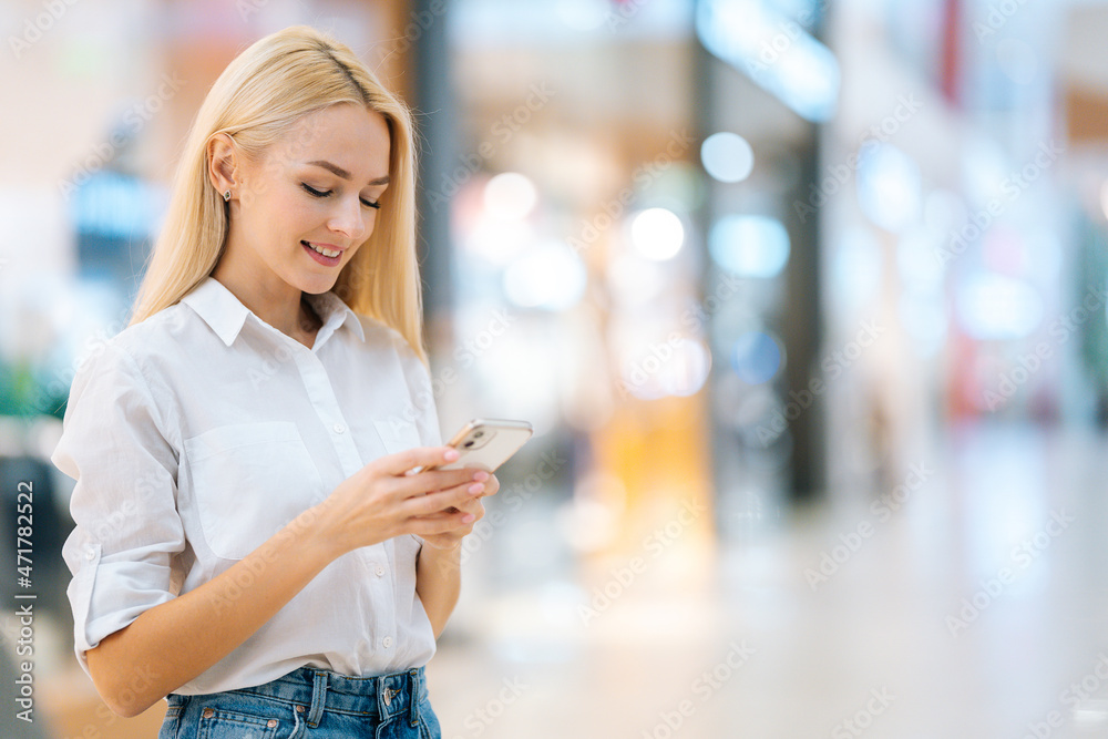 Portrait of smiling attractive blonde young woman with long hair in stylish clothing using mobile phone, standing in hall of mall centre, blurred background, looking at smartphone screen. Portrait of smiling attractive blonde young woman with long hair in stylish clothing using mobile phone, standing in hall of mall centre, blurred background, looking at smartphone screen.