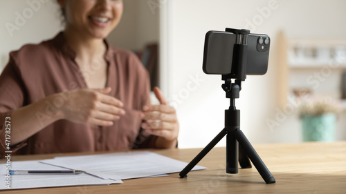 Focus on cellphone standing on table on tripod stabilizer, motivated indian young female blogger recording video or stories on smartphone web camera, talking streaming online, videoblog concept.