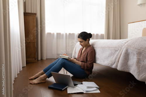 Happy young indian woman sitting on warm wooden floor in cozy bedroom, preparing for professional examination, enjoying studying on online courses, writing notes in copybook, educational concept.