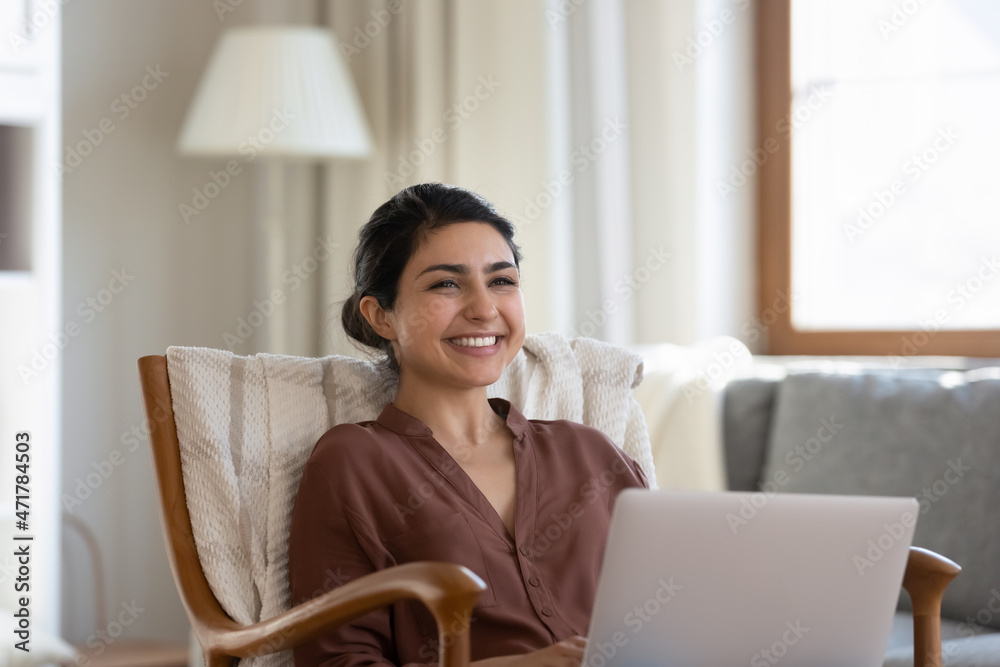 Distracted from computer work smiling millennial indian ethnicity woman looking in distance, sitting in cozy armchair, feeling excited of getting message with good news, daydreaming resting at home. Distracted from computer work smiling millennial indian ethnicity woman looking in distance, sitting in cozy armchair, feeling excited of getting message with good news, daydreaming resting at home.