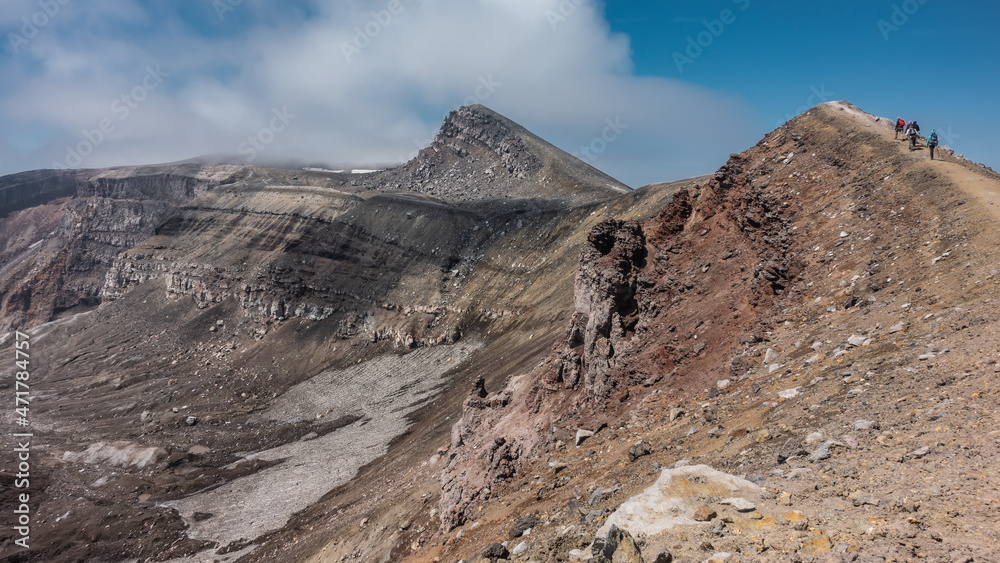 The trail runs along the edge of a volcanic crater. The layered structure of the soil. Tiny silhouettes of climbing tourists are visible on the path. Blue sky, clouds. Kamchatka. Gorely Volcano