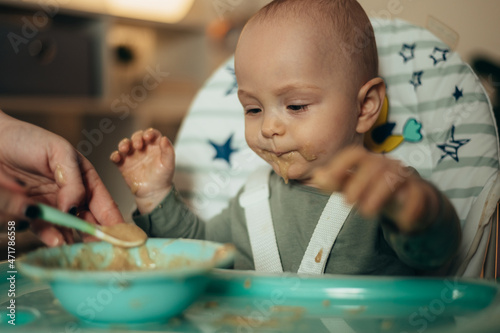 Baby eating with a face stained in food while mother feeds him with a spoon