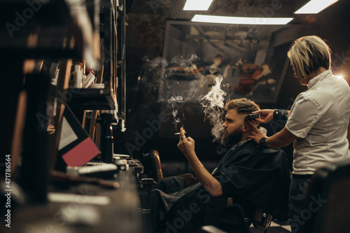 Young bearded man getting haircut by hairdresser and smoking a cigarette